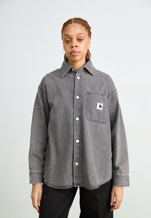 Camicetta in denim grigio con colletto, grande tasca frontale e bottoni bianchi. Tessuto morbido con una vestibilità rilassata e dettagli di cuciture a contrasto.