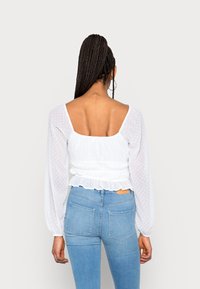 Kvinna med långt flätat hår klädd i en vit genomskinlig långärmad topp och ljusblå högmidjade jeans, vänd bort.