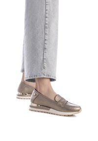 Zapatos metálicos dorados tipo slip-on con un acabado texturizado, que presentan una correa en la parte frontal y un tacón bajo con un acento plateado. Jeans grises visibles.