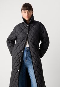 Svart, quiltet lang frakk med høy krage, trykknapper og strukturert overflate, paret med blå denimjeans.