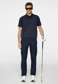 Mörkblå pikétröja med korta ärmar och ribbad nederkant, kombinerad med mörkblå cargobyxor. Modellen har solglasögon och håller en golfklubba.