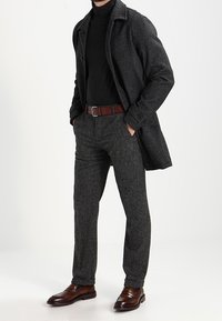 Grauer Wollmantel über einem schwarzen Rollkragenpullover, kombiniert mit grauen strukturierten Hosen und braunen Lederschuhen. Ein brauner Gürtel rundet das Outfit ab.