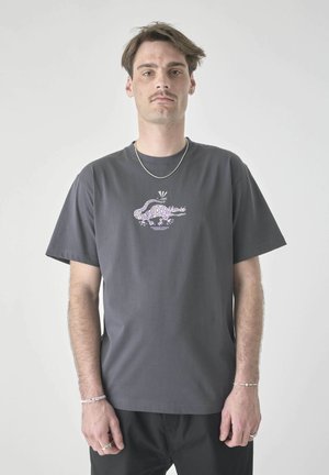 Man with mustache wearing gray T-shirt with purple patterned crocodile graphic and text, standing against plain light gray background.