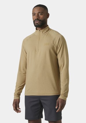 Hombre con sudadera beige de manga larga y cremallera corta y pantalones cortos gris oscuro, de pie contra un fondo claro y liso.