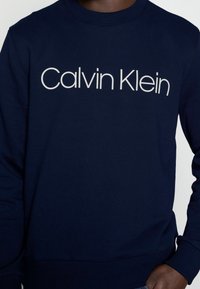 Granatowa bluza wykonana z bawełnianej mieszanki, z prążkowanymi mankietami i dołem. Posiada duże białe logo "Calvin Klein" w czcionce bezszeryfowej.