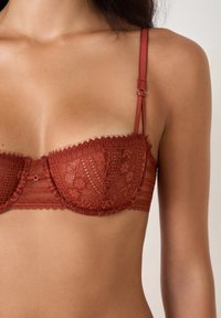 Reggiseno in pizzo ruggine con bordi scallop, supporto con ferretto, spalline in satin regolabili e un motivo floreale su uno sfondo di caldo colore ruggine.