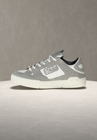 Γκρι και λευκό σουέντ αθλητικό παπούτσι με λευκά κορδόνια, ελαστική σόλα και λογότυπο "GCDS" στο πλάι και στη φτέρνα, τοποθετημένο σε ανοιχτόχρωμη επιφάνεια.