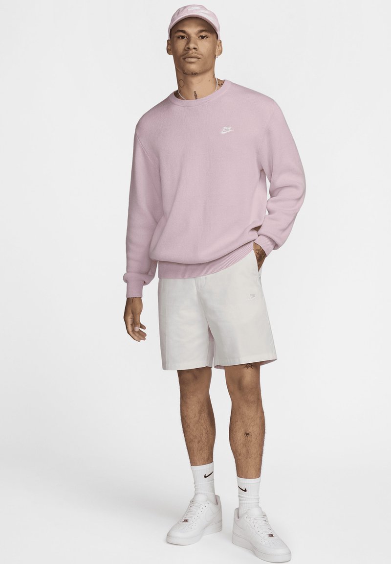 Jasno różowy sweter z dzianiny z okrągłym dekoltem, w połączeniu z białymi szortami. Outfit uzupełnia pasująca różowa czapka i białe trampki.