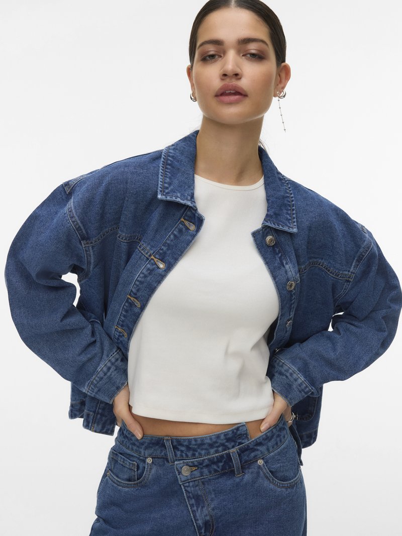 Young woman with tied-back hair wearing a white cropped top, oversized denim jacket, and unbuttoned blue jeans against a white background.