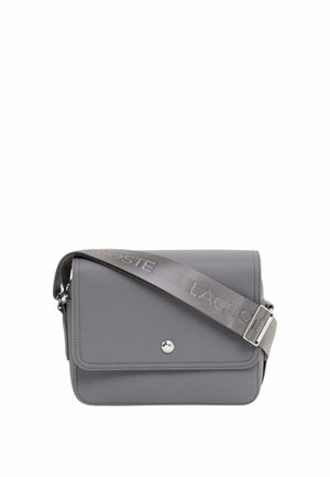 Sac bandoulière gris avec rabat avant, fermeture par bouton argenté, et large sangle réglable embossée du logo "Lacoste".