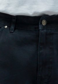 Μαύρα τζιν παντελόνια με κανονική γραμμή, που διαθέτουν μεταλλικό κλείσιμο κουμπιού και ορατές ραφές. Το ύφασμα έχει λεία υφή.