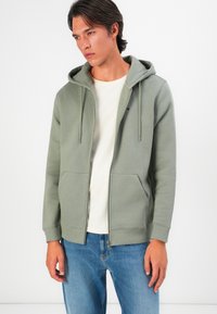 Joven usando una sudadera ligera verde claro con cremallera sobre una camiseta blanca y jeans azules, de pie contra un fondo blanco liso.