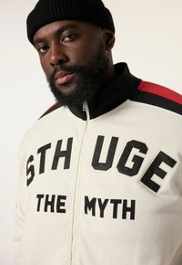 Giacca leggera con zip di colore chiaro, con bold lettering nero "STHUGE THE MYTH", accentuata da strisce rosse e nere sulle spalle. Materiale morbido, vestibilità rilassata.