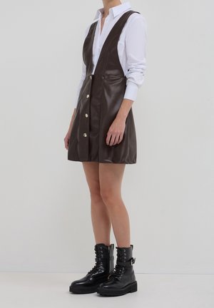 Osoba ubrana w brązową, bezrękawną, skórzaną sukienkę zapinaną na guziki, na białą koszulę z kołnierzykiem, zestawioną z czarnymi, sznurowanymi botkami za kostkę.