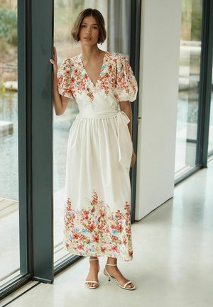 Woman in white floral wrap dress with puffed sleeves and ankle-strap heels standing by glass door in modern bright room.