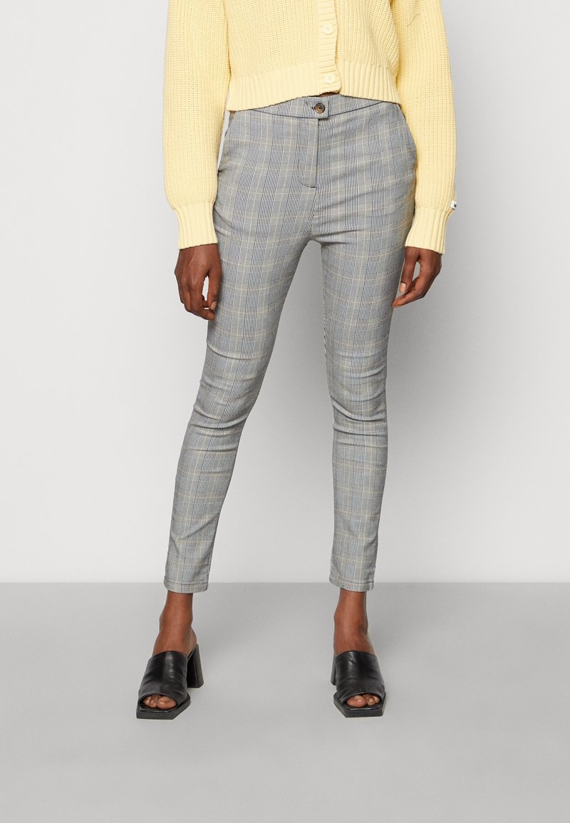 Personne portant un pantalon ajusté à carreaux gris, un pull court en maille jaune et des chaussures noires à talons carrés ouverts, debout sur un sol gris.