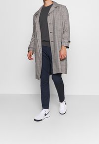 Grau karierten Trenchcoat mit Knopfverschluss, getragen über einem dunkelgrauen Hemd, Marineblauen Hosen und weißen Sneakers mit schwarzen Akzenten.