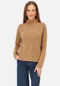 Beżowy sweter dziergany o wysokim dekolcie, z teksturowanym wzorem diamentowym oraz ściągaczami na mankietach i u dołu; zestawiony z ciemnymi jeansami.