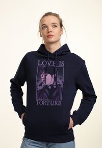 Joven mujer con sudadera azul marino con un gráfico púrpura de una chica y el texto "EL AMOR ES TORTURA", de pie con las manos en los bolsillos mirando hacia arriba.