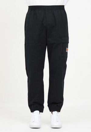 Pantalon cargo noir avec taille et poignets élastiques, poche latérale avec rabat, porté avec des baskets blanches, visible de la taille aux pieds.