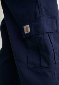 Pantaloni cargo de culoare bleumarin, confecționați dintr-un material durabil, care au multiple buzunare și o etichetă cusută cu un logo galben pe lateral.