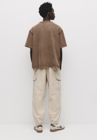Bruine oversized korte mouw shirt, beige cargobroek met elastische boorden en sneakers in neutrale kleuren. Matte textuur, ontspannen pasvorm en functionele zakken.