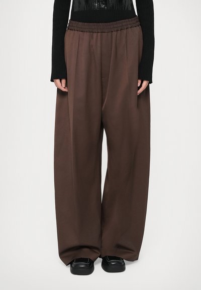 Persona que lleva pantalones anchos marrones de pierna ancha con cintura elástica y zapatos negros, combinados con una camiseta negra de manga larga.