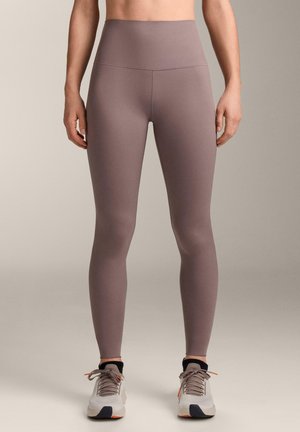 Ženska oblečena v visokoločene vijoličaste legice in bele tekaške superge, stoji na nevtralni enobarvni podlagi.