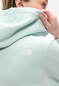 Žena si upravuje svetlomodrú mikinu North Face, prezentujúc logo na zadnom ramene na bielom pozadí.