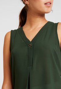 Blusa verde sin mangas con escote en V, detalle de pliegues y un botón dorado como adorno. Confeccionada con una tela suave y ligera.