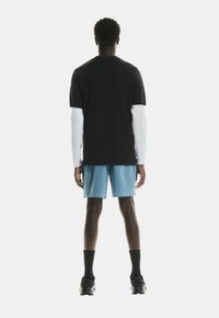 Camisa negra de manga corta sobre mangas blancas largas, combinada con pantalones cortos azul claro y zapatillas deportivas negras, vista desde atrás.