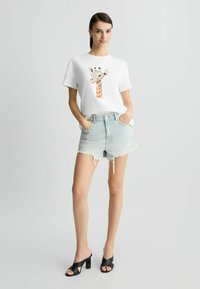 T-shirt blanc avec un imprimé girafe, associé à un short en denim léger bleu déchiré. Des sandales à talons noires complètent la tenue.