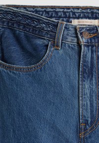 Jean en denim bleu en gros plan montrant le détail de la ceinture tressée, la poche avant, le bouton et l’étiquette indiquant « Ribcage Wide Leg ».