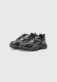 Schwarze Sportschuhe mit einem strukturierten Mesh-Obermaterial, silbernen Akzenten, runder Zehenpartie, dicke Gummisohle und auffälligem Schnürsystem.