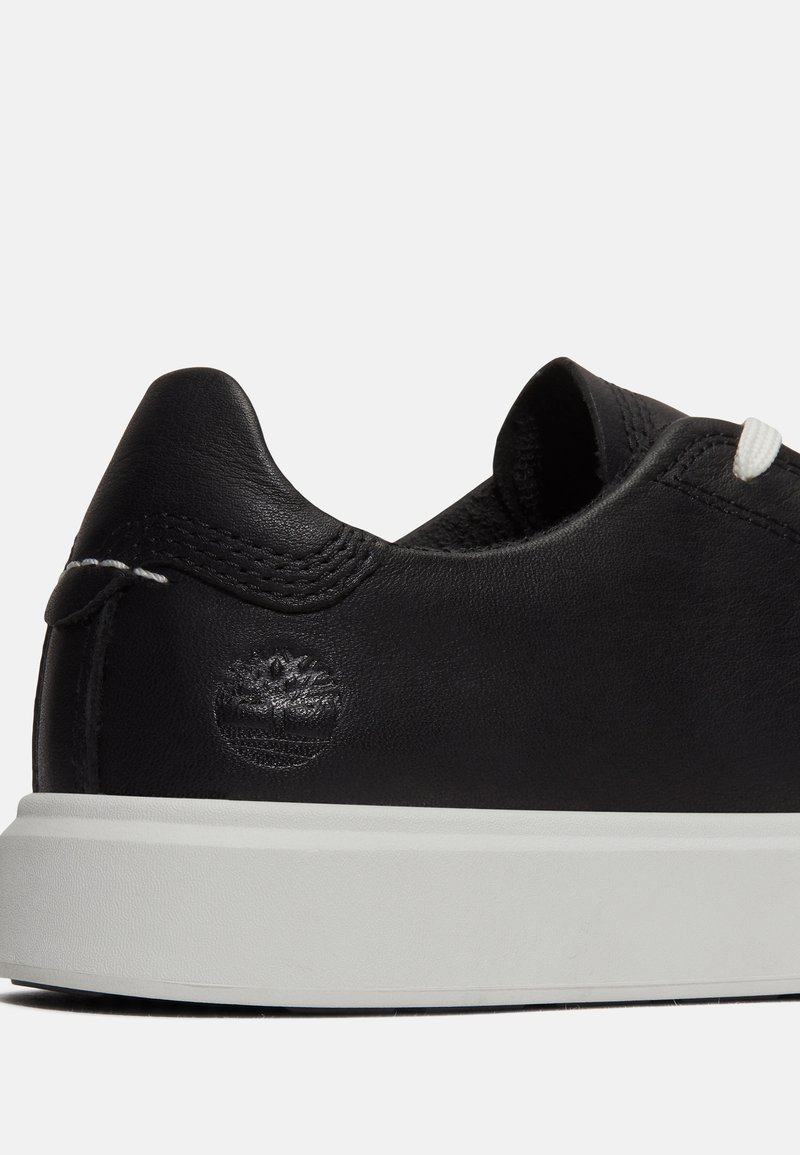 Zapatilla negra de cuero con suela de goma blanca, que presenta un diseño cosido y un logo estampado en el talón. Cierre con cordones de color contrastante.