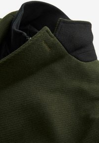 Grön zip-up jacka med en strukturerad, fleece-liknande yta och en svart, slät krage. Har en diskret logotagg.