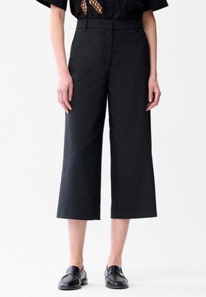 Persoon draagt wijde, verkorte zwarte broek en zwarte loafers, getoond van taille tot voeten, staand tegen een effen witte achtergrond.