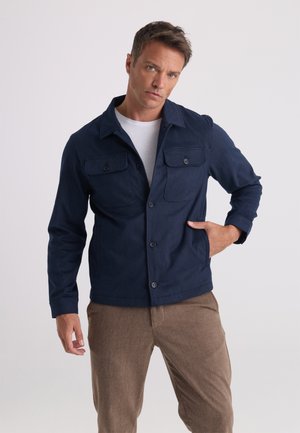 Uomo che indossa una giacca blu navy con bottoni e tasche con patta sul petto e pantaloni marroni, in piedi con una mano nella tasca.