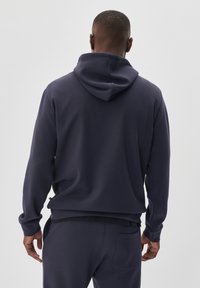Sudadera azul marino con capucha ajustable, textura suave, corte relajado y detalles de bolsillo lateral, vista desde la parte de atrás con pantalones a juego.