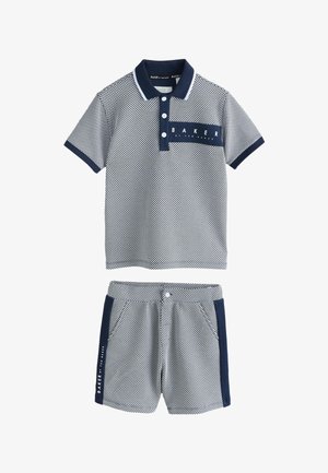 Unge dreng smilende, iført kortærmet mønstret poloshirt og matchende shorts med sidelister, hænderne i lommerne, stående mod en ensfarvet baggrund.