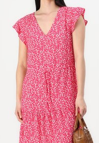 Rosa Blumenkleid mit V-Ausschnitt, kurzen, flatternden Ärmeln, gestuftem Rüschensaum und leichtem Stoff. Verziert mit kleinen weißen Blumen.