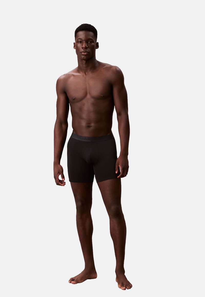 Czarne bokserki wykonane z miękkiego materiału, o dopasowanym kroju i elastycznym pasie. Model stoi boso na jasnym tle.