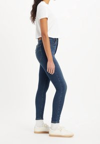 Jeans ajustados de denim azul oscuro, de cintura alta, con un bolsillo trasero y costuras mínimas. Combinados con zapatillas blancas y calcetines hasta el tobillo.