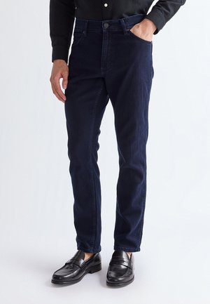 Bărbat purtând blugi slim fit de culoare albastru închis, pantofi loafer din piele neagră și o cămașă neagră cu nasturi, cu o mână în buzunar.