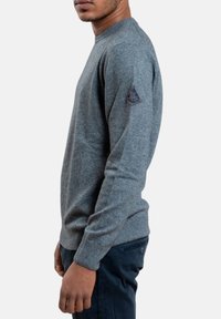 Maglione grigio in tessuto a maglia liscia con scollatura rotonda e polsini a coste. Presenta una toppa a forma di diamante sulla spalla sinistra.