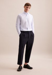 Camicetta a righe azzurro chiaro con colletto button-down, abbinata a pantaloni navy plissettati e scarpe nere con lacci, in piedi su uno sfondo neutro.