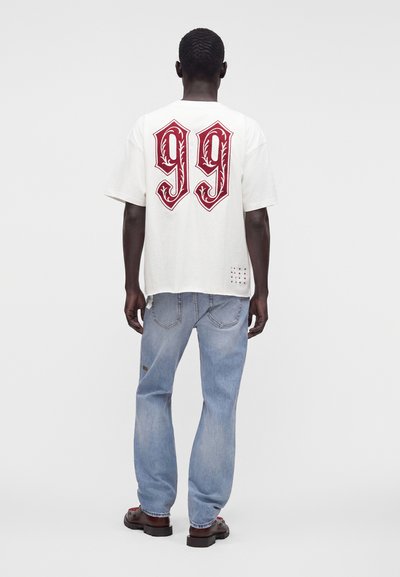 Άνδρας με σκούρο δέρμα που φοράει λευκό T-shirt με μεγάλο κόκκινο "99" στην πλάτη, ανοιχτό μπλε τζιν και σκούρα παπούτσια, στέκεται με την πλάτη προς τα εμπρός.