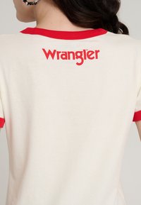 T-shirt en coton blanc avec col et manches côtelés rouges, arborant le logo "Wrangler" en rouge vif au dos. Texture lisse et ajustée.