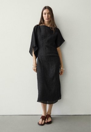 Kobieta o długich brązowych włosach, ubrana w czarną, teksturowaną, midi sukienkę z szerokimi rękawami oraz brązowe sandały z odkrytymi palcami, stojąca na tle jednolitej jasnej ściany.