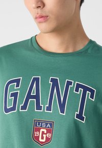 Zelené bavlněné tričko s velkým potiskem "GANT" v námořnické modři, s logem USA ve štítu v červené a zlaté barvě na spodním okraji. Klasický design s kulatým výstřihem.